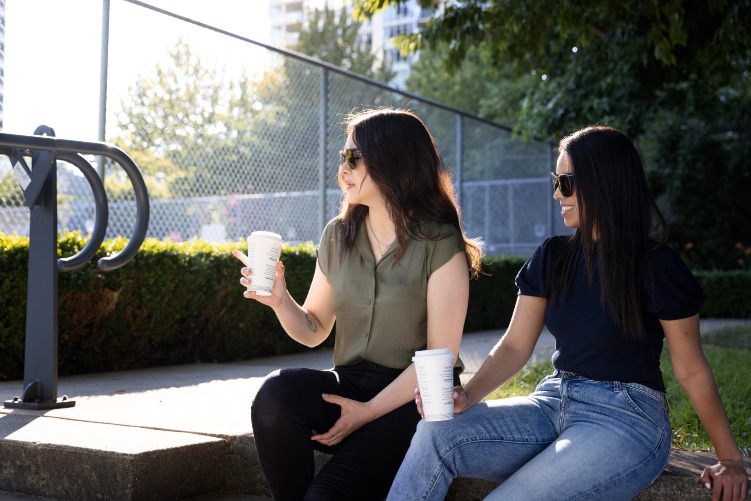  two friends laughing on a park bench with coffee cups, late afternoon light