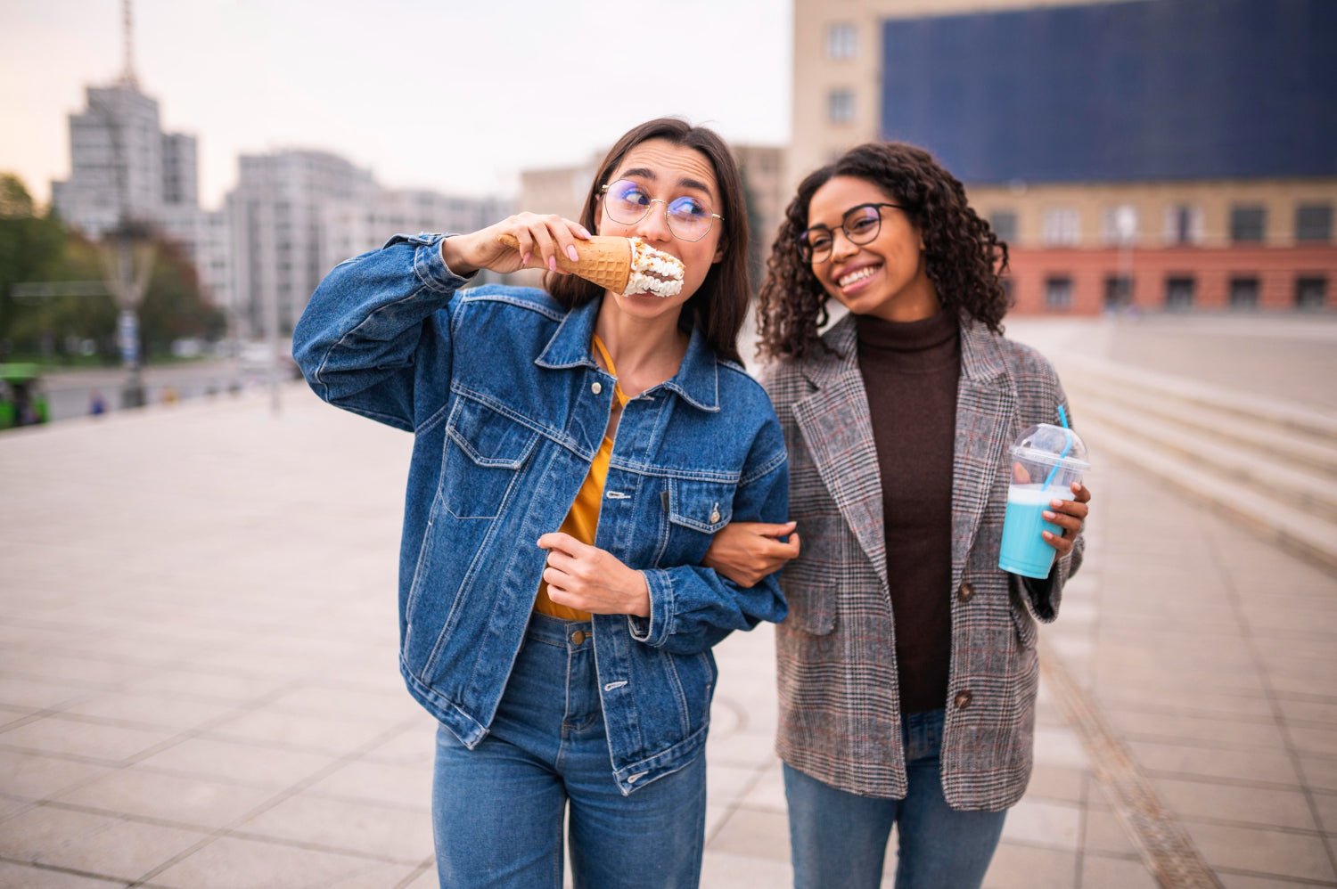 two friends laughing while walking with iced coffees on a sunny sidewalk